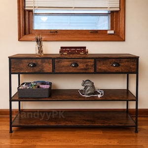 May include: A rectangular console table with a dark brown wood grain finish and a black metal frame. It features three top drawers with black semi-circular pulls, two open lower shelves, a glass jar with sticks, a wooden box, and an elephant figurine.