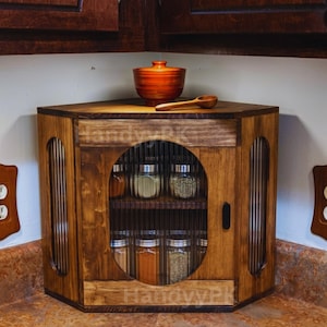 May include: A wooden corner spice rack with a round door and glass jars filled with spices. The rack is stained a warm brown color and has a small wooden bowl and spoon on top. The text "HandywrkPk" is visible.