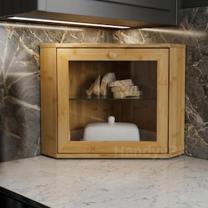May include: A corner cabinet made of light-colored bamboo with a clear glass front door and a single knob. Inside, a glass shelf holds several slices of bread, while the bottom shelf contains a white, rectangular, textured butter dish.
