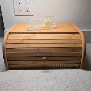 May include: A natural bamboo bread box with a roll-top design and a small wooden knob. A clear, rectangular glass butter dish sits on top. The bread box is made of light brown bamboo and has a smooth finish.