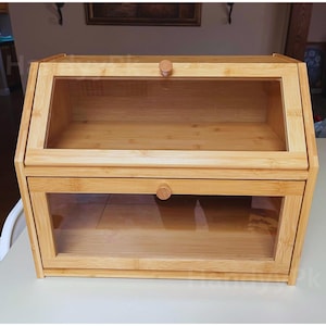 May include: A light brown bamboo bread box with a clear glass front. The bread box has a hinged top and a pull-down door, both with small wooden knobs. The interior is also made of bamboo.