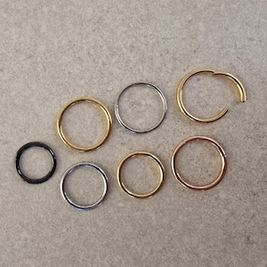 May include: A selection of seven circular body jewellery items in different metallic finishes. The collection includes black, silver, gold, and rose gold tones. One gold piece is open, with a hinged design. The jewellery is displayed on a neutral grey surface.