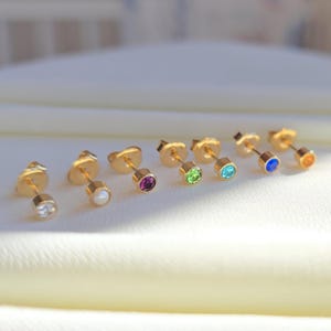 May include: A collection of gold-colored stud earrings with various colored gemstones. The earrings are arranged in a row, showcasing a range of colors including clear, white, purple, green, blue, and orange. The studs are small and delicate.