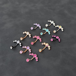 May include: A collection of curved barbell body jewelry pieces, each featuring a row of four circular gemstones in various colors including clear, pink, blue, and iridescent, set in metallic bands of silver, gold, and black tones.