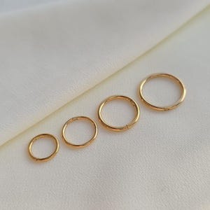 Könnte beinhalten: Vier kreisförmige goldfarbene Creolen, die der Größe nach von klein nach groß in einer Reihe angeordnet sind. Jeder Ring hat eine glatte, polierte Oberfläche und ein nahtloses, durchgehendes kreisförmiges Banddesign in schlichter Optik.
