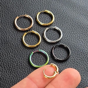 Könnte beinhalten: Eine Auswahl an Creolen in verschiedenen Metallfarben, darunter Gold, Silber und Schwarz. Ein einzelner Ohrring ist geöffnet und zeigt den Verschluss. Die Ohrringe liegen auf einer strukturierten, dunklen Oberfläche.