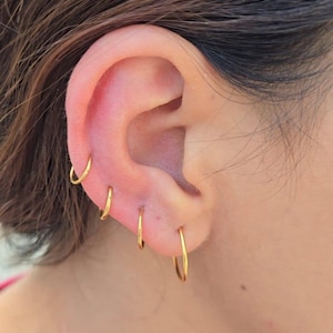 May include: A set of four thin, polished gold hoop earrings arranged in a vertical line along the earlobe and cartilage. Each earring is a simple, circular wire hoop with a smooth, reflective metallic finish, creating a uniform look.