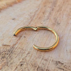 May include: A gold-coloured, hinged hoop earring, open and slightly curved. The earring is made of a shiny metal and is set against a light brown, textured wooden surface.