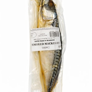 May include: A whole smoked mackerel in a transparent plastic wrapper. The fish has a silver and black striped pattern. The packaging includes a label with the text "SMOKED MACKEREL".