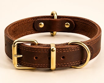 Puede incluir: Un collar para mascota de cuero marrón oscuro con herrajes de metal en tono dorado, que incluye una hebilla resistente, una anilla en D para la correa y una trabilla fija. El collar presenta costuras visibles en los bordes y dos remaches.