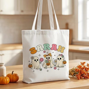 Peut inclure: Un sac fourre-tout en toile blanche orné d'un motif coloré représentant trois fantômes tenant des citrouilles et des fleurs, avec le nom SARAH imprimé en grandes lettres ondulées multicolores au-dessus, agrémenté de petites étoiles.