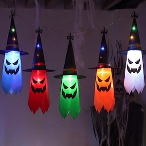 Könnte beinhalten: Eine Lichterkette mit fünf hängenden Geisterdekorationen, die jeweils einen schwarzen spitzen Hexenhut mit farbigem Band tragen. Die Geister leuchten in Blau, Rot, Grün, Orange und Weiß und haben aufgedruckte schwarze Gesichter mit gezackten Augen und Mündern.