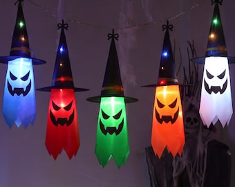 May include: A string of five hanging ghost decorations, each wearing a black pointed witch hat with a colored band. The ghosts are illuminated in blue, red, green, orange, and white, featuring black printed facial expressions with jagged eyes and mouths.