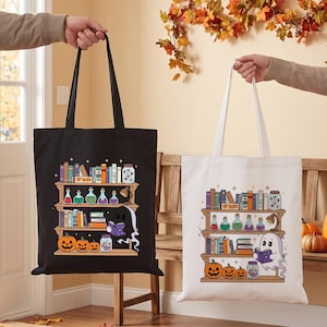 Op de afbeelding: Twee tote bags, een zwarte en een witte, elk met een grafische print van een houten boekenplank. De planken bevatten kleurrijke boeken, toverdrankflesjes, een lamp, een pot met oogballen en drie pompoenen. Een klein spookje leest een boek.