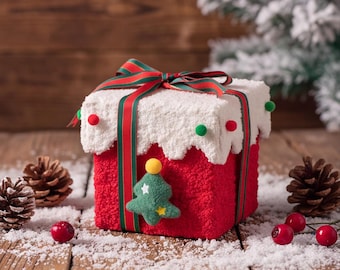 May include: A square red gift box with a textured surface, topped with a white scalloped lid resembling snow. The box is wrapped in a green and red striped ribbon and decorated with small red and green pom-poms and a felt Christmas tree.