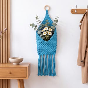May include: A bright blue macrame wall hanging pocket featuring a diamond-shaped woven pattern, suspended from a circular wooden ring. The pocket contains a bouquet of white daisies with yellow centers and green eucalyptus leaves, finished with long tassels.