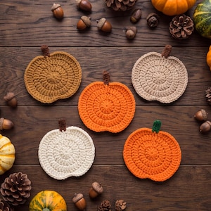May include: Five circular crochet coasters shaped like pumpkins in shades of mustard yellow, beige, bright orange, cream, and pumpkin orange. Each coaster features a small brown or green crocheted stem at the top and a textured spiral stitch pattern.
