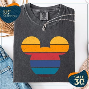 a gray t-shirt with a colorful Mickey Mouse design, along with a pair of jeans and a beige sweater. The t-shirt is displayed on a white background, and there are two sale signs indicating a 30% discount.
