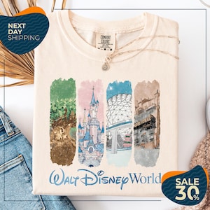 A white t-shirt with a graphic design featuring illustrations of various Disney landmarks, including the castle, the Eiffel Tower, and the Hollywood sign. The t-shirt is displayed on a white background, and there are jeans and a sweater visible in the background.