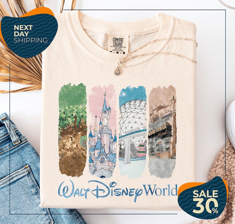 A white t-shirt with a graphic design featuring illustrations of various Disney landmarks, including the castle, the Eiffel Tower, and the Hollywood sign. The t-shirt is displayed on a white background, and there are jeans and a sweater visible in the background.