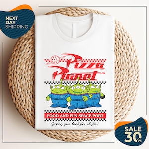 A white t-shirt with a graphic design featuring three alien characters from the Pixar movie "Pizza Planet" and the text "Food and Fun Space Port" along with a sale offer.