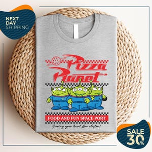 a gray t-shirt with a graphic design of three alien characters from the Pixar movie "Pizza Planet" and text promoting a food and fun space port.