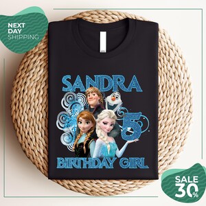 A black t-shirt with a design featuring characters from the Disney movie Frozen, specifically Elsa, Olaf, and Anna. The text on the t-shirt reads "SANDRA BIRTHDAY GIRL".