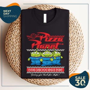 a black t-shirt with a graphic design of three alien characters from the animated film "Pizza Planet" and text promoting a food and fun space port.
