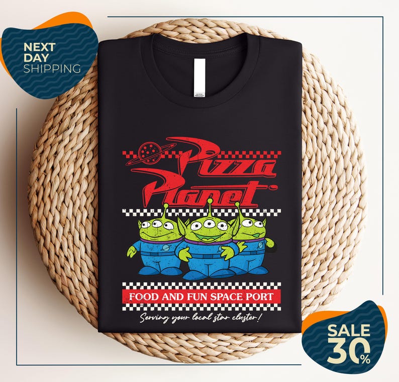 a black t-shirt with a graphic design of three alien characters from the animated film "Pizza Planet" and text promoting a food and fun space port.