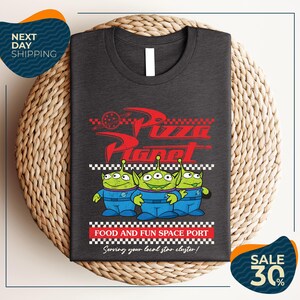 a gray t-shirt with a graphic design of three alien characters from the Pixar movie "Pizza Planet" and text promoting a food and fun space port.