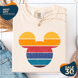 a white t-shirt with a colorful Mickey Mouse design, along with a pair of blue jeans and a beige sweater. The t-shirt is displayed on a white background, and there are two sale signs indicating a 30% discount.