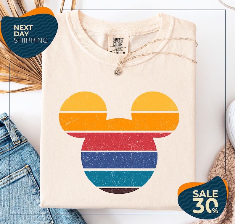 a white t-shirt with a colorful Mickey Mouse design, along with a pair of blue jeans and a beige sweater. The t-shirt is displayed on a white background, and there are two sale signs indicating a 30% discount.