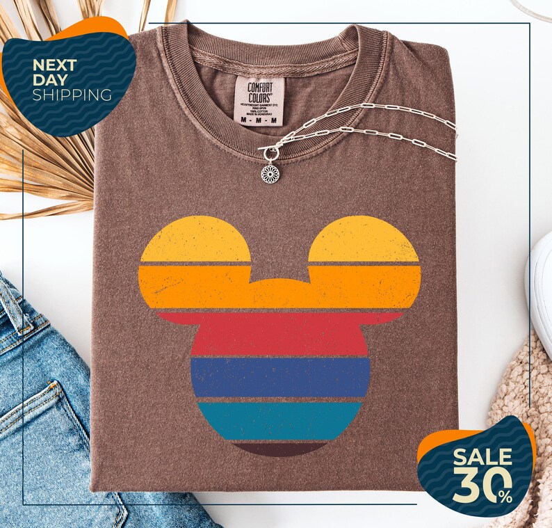 a brown t-shirt with a colorful Mickey Mouse design, along with a denim jacket and a necklace. The t-shirt is displayed on a white background, and there are two sale signs indicating a 30% discount.