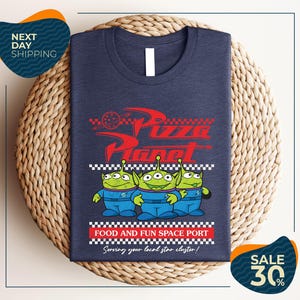 A t-shirt with a graphic design featuring three alien characters from the Pixar movie "Pizza Planet" and the text "Food and Fun Space Port" along with a sale offer.