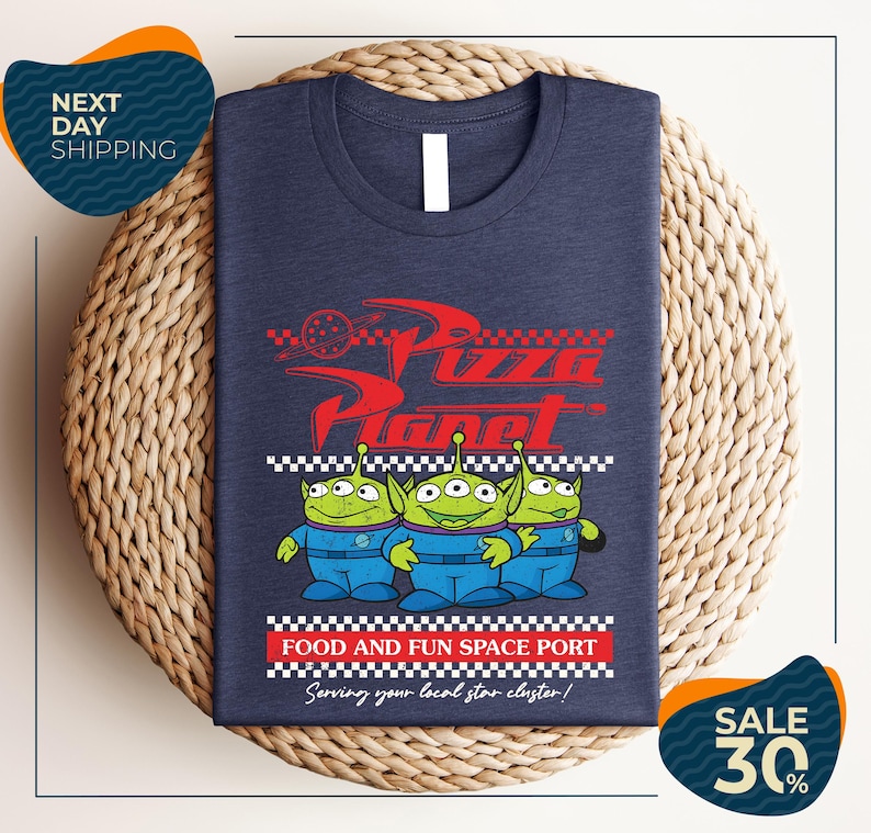 A t-shirt with a graphic design featuring three alien characters from the Pixar movie "Pizza Planet" and the text "Food and Fun Space Port" along with a sale offer.