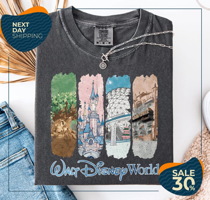 a gray t-shirt with a graphic design depicting various Disney World landmarks, including the Walt Disney World resort, the Wood Tower, and the Epcot Pavilion. The t-shirt is displayed on a white background, and there are jeans and a necklace visible in the foreground.