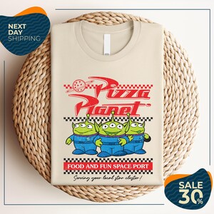 a t-shirt with a design related to the popular animated movie "Pizza Planet". The t-shirt is displayed on a woven basket, and there are two sale signs in the image.