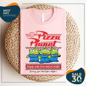 A pink t-shirt with a graphic design featuring three alien characters from the Pixar movie "Pizza Planet" and the text "Food and Fun Space Port" along with a sale offer.