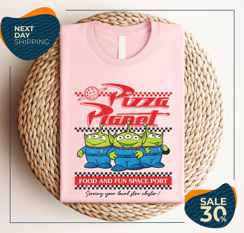 A pink t-shirt with a graphic design featuring three alien characters from the Pixar movie "Pizza Planet" and the text "Food and Fun Space Port" along with a sale offer.