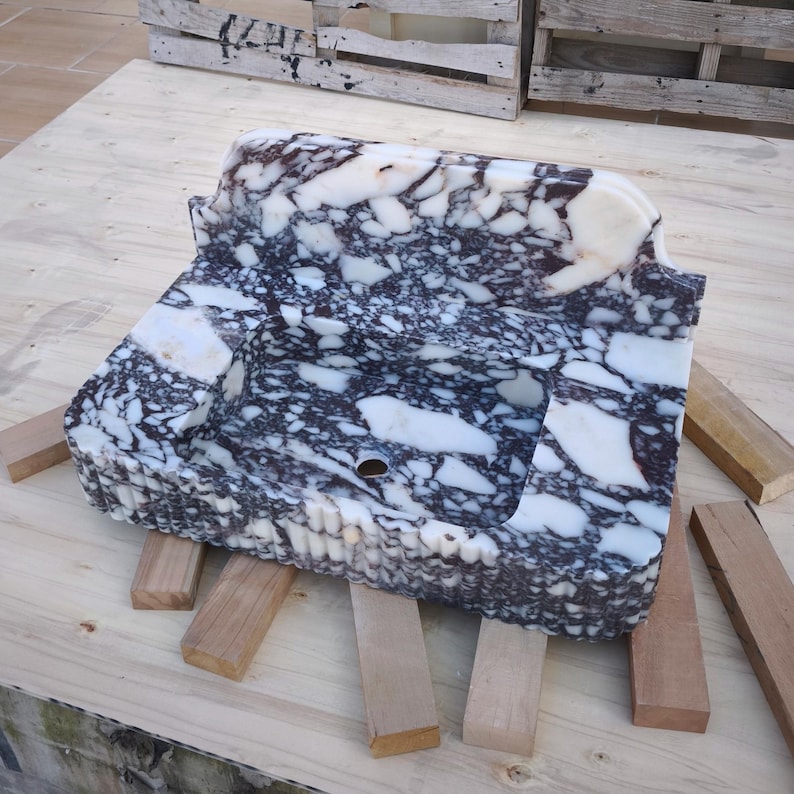 May include: A rectangular marble sink with a white and dark gray pattern. The sink has a curved back and a small hole near the front. It rests on several small wooden supports, with a wooden pallet in the background.