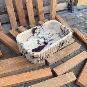 Op de afbeelding: Rechthoekige marmeren wastafel met een gegroefde buitenkant. De wastafel heeft een witte basis met donkerbruine en bordeauxkleurige adering. De wastafel staat op houten planken.