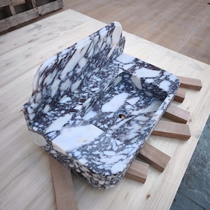 May include: A rectangular marble sink with a white and dark gray veined pattern. The sink has a raised back and a basin with a drain hole. It is supported by wooden blocks on a light wood surface.