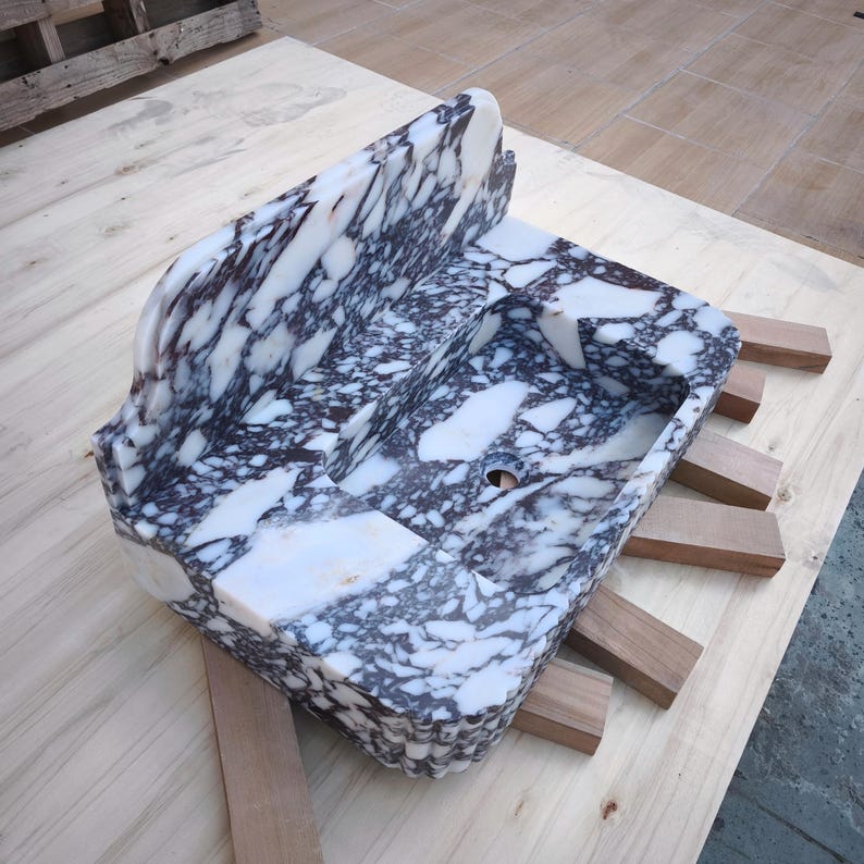 May include: A rectangular marble sink with a white and dark gray veined pattern. The sink has a raised back and a basin with a drain hole. It is supported by wooden blocks on a light wood surface.
