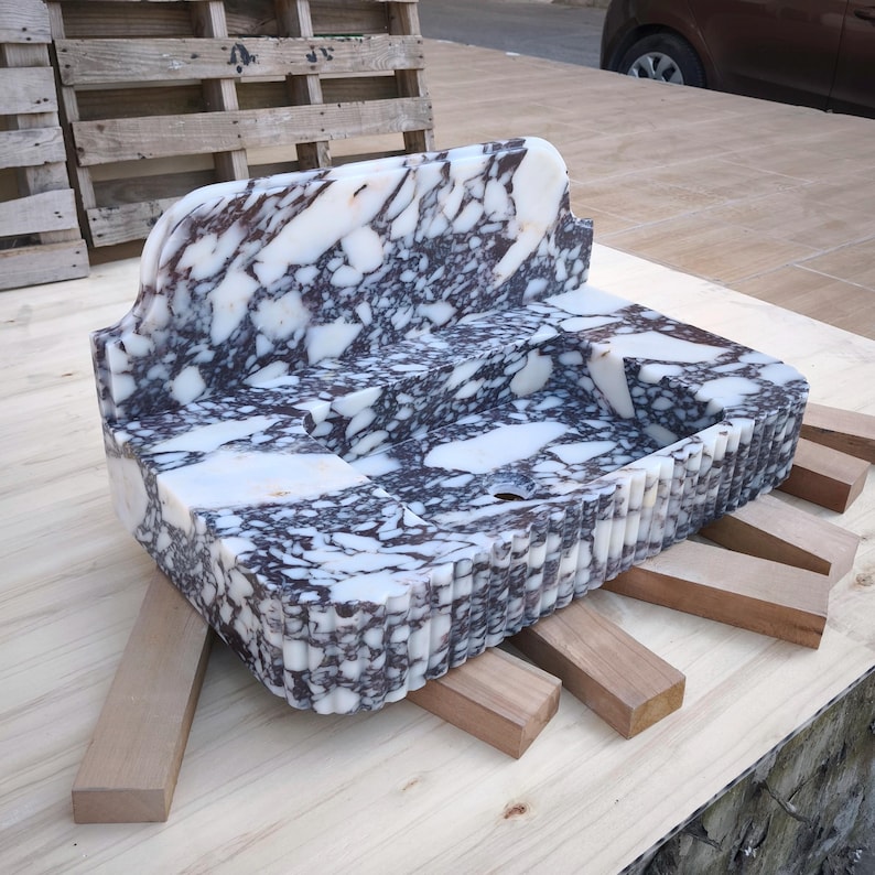 May include: A rectangular marble sink with a dark purple and white pattern. The sink has a curved back and a fluted edge. It is supported by wooden beams and sits on a light-colored surface.