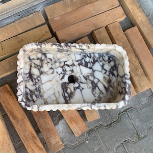 Op de afbeelding: Rechthoekige marmeren wastafel met een geschulpte rand. De wastafel heeft een wit en bruin geaderd patroon en een centraal afvoergat. De wastafel staat op een houten pallet.