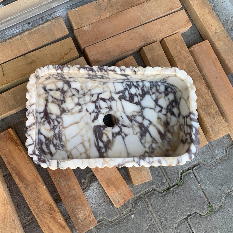 Op de afbeelding: Rechthoekige marmeren wastafel met een geschulpte rand. De wastafel heeft een wit en bruin geaderd patroon en een centraal afvoergat. De wastafel staat op een houten pallet.