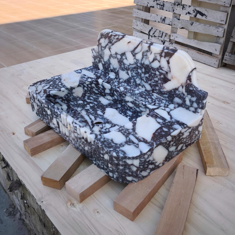 May include: A large, carved stone sink with a unique shape and a striking black and white speckled pattern. The sink is supported by wooden beams and sits on a wooden surface. The stone has a polished finish.