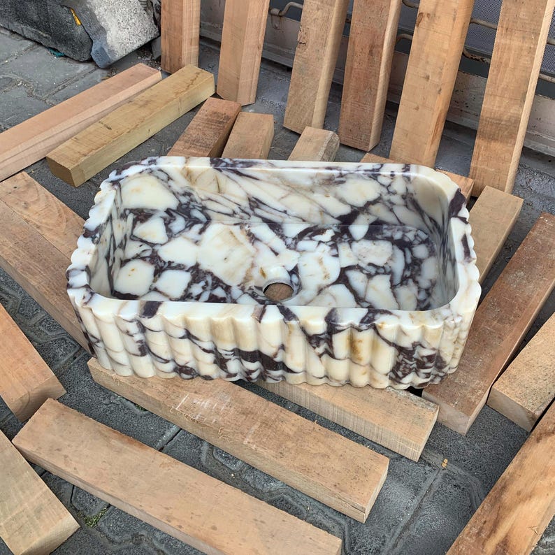 Op de afbeelding: Een rechthoekige marmeren wastafel met een cr&egrave;me- en donkerbruin patroon. De wastafel heeft een gegolfde rand en een centraal afvoergat. Hij rust op houten planken.