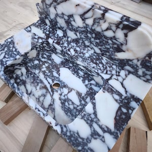 May include: A rectangular marble sink with a unique pattern of white and dark gray veining. The sink has a curved back and a rounded basin with a drain hole. The edges are scalloped.