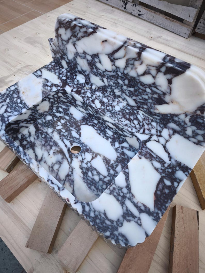 May include: A rectangular marble sink with a unique pattern of white and dark gray veining. The sink has a curved back and a rounded basin with a drain hole. The edges are scalloped.